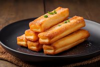 Fresh baked churros stacked on black plate with sugar coating and green herb garnish, served on rustic wooden table with brown textile napkin.