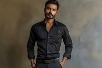 South Asian man in black button-up shirt standing confidently against textured gray wall, perfect for professional portraits or fashion advertising.