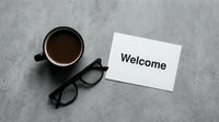 Welcome card with coffee cup and glasses on gray concrete surface, flat lay composition for business greeting concepts.