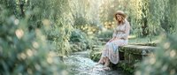 Young woman in floral dress and hat sitting on stone bridge in peaceful garden setting with weeping willow trees and natural lighting for lifestyle content.