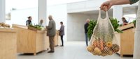 Hand holding mesh shopping bag with fresh produce in modern grocery store with customers and wooden checkout counters in background for retail and sustainable shopping concepts.