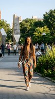 Young African American woman with dreadlocks walking confidently through urban park plaza wearing colorful patterned outfit during golden hour sunset.