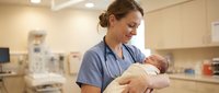 Female healthcare professional in scrubs holding newborn baby in modern hospital nursery setting with medical equipment and warm lighting.