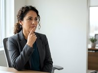 Mature woman with curly dark hair and olive complexion wearing charcoal blazer, seated at desk with hand to chin in thoughtful pose, contemplating business decision in bright office environment.
