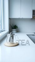 Glass teapot on wooden trivet beside a wire TEA sign on a white kitchen counter, minimalist Scandinavian interior with bright natural light.