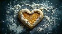 Heart-shaped homemade pastry with apricot jam filling on dark baking surface dusted with flour, showcasing rustic baking and love-themed food preparation.
