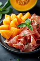 Fresh cantaloupe melon slices with prosciutto ham on dark plate garnished with basil leaves for gourmet appetizer and Mediterranean cuisine presentation.
