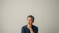 Middle-aged Asian male professional in dark blue casual shirt, hand thoughtfully positioned near chin, contemplative expression, neutral studio background, suitable for business, corporate, and lifestyle editorial use.
