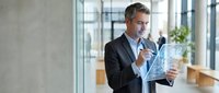 Middle-aged Caucasian male professional businessman in formal black suit and light blue shirt holding digital tablet with holographic data visualization in modern office environment.