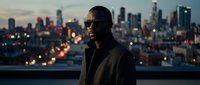 African American male in dark sunglasses and charcoal jacket stands against illuminated city skyline at dusk, urban professional portrait for business and lifestyle content.