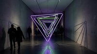 Neon glowing triangles installation in a dark corridor with silhouetted visitors, purple and green lights reflecting on concrete walls and floor.