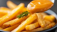 Golden french fries on black plate with rosemary garnish and dripping sauce. Close-up macro shot shows crispy potato texture and glistening surface in warm lighting.