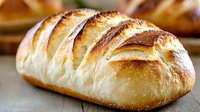 Fresh baked white bread loaf with golden brown crust and distinctive scoring pattern on wooden surface, close up view showing soft texture and artisanal quality.