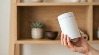 Young adult hand holding blank white cylindrical container against wooden shelving with potted plants and home decor items for product packaging mockup.