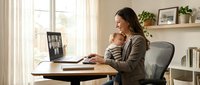 Young mother multitasking while working from home office, holding baby during video conference call on laptop computer in bright modern workspace.