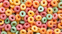 Colorful fruit-flavored cereal rings in pink, yellow, and green create vibrant breakfast food pattern. Close up view of sweet round loops shows texture and detail.