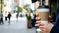 Person holding takeaway coffee cup on urban street with blurred pedestrians and buildings in background, suitable for beverage, lifestyle, and commercial use.