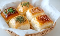 Fresh baked dinner rolls with golden brown crust topped with sesame seeds and herbs served in wicker basket on white paper, close up view of soft bread.