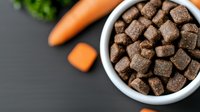 Dry pet food kibbles in white ceramic bowl with fresh carrots on dark background, top view. Healthy natural dog or cat nutrition concept.