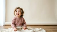 Toddler with curly blonde hair, Caucasian, wearing pink sweater, sitting on knitted blanket, smiling joyfully indoors for parenting, childcare, family lifestyle content.