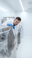 Male scientist in white lab coat and safety glasses working with laboratory equipment in sterile research facility for pharmaceutical development.