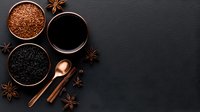 Dark moody composition with coffee grounds, black tea, cinnamon sticks and star anise on black stone background, copper spoon adds warmth to beverage ingredients.