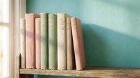 Row of vintage books in pastel colors on wooden shelf against mint green wall with natural window light creating soft shadows for cozy reading nook decor.