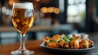 Glass of golden beer with bubbles alongside plate of glazed chicken wings garnished with fresh herbs on wooden table in restaurant setting with blurred bokeh background.