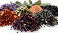 Colorful piles of loose tea leaves and spice powders arranged on white surface, showing variety of natural herbal ingredients and textures in warm earthy tones.