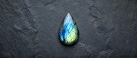 Labradorite gemstone with blue and green flash on dark textured slate surface. Natural mineral specimen displaying iridescent colors for jewelry design.