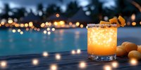 Glowing orange cocktail with salted rim and citrus garnish on wooden deck by illuminated pool at dusk, resort lights creating bokeh effect in background.
