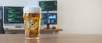 Cold beer glass with foam on wooden desk in modern office workspace with computer monitors displaying code and data analytics in background for relaxation concept.