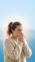 Young woman with light brown hair pulled back in a ponytail, Caucasian ethnicity, wearing a cream knit sweater, sneezing or blowing nose into tissue against blue gradient background.