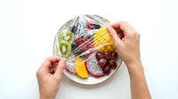 Hands wrapping plastic film around plate with fresh fruit assortment including kiwi, dragon fruit, and grapes for food storage and preservation.
