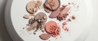 Broken makeup powder compacts in warm nude and coral tones scattered on white surface creating artistic beauty product flatlay composition for cosmetics branding.