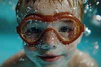 Young child wearing orange swimming goggles underwater in pool with bubbles and water droplets creating playful summer atmosphere, close up portrait with bright blue water.