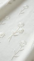 White cotton fabric with delicate embroidered daisy flowers and stems creating elegant floral pattern for clothing and home textile projects.