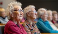 Senior Caucasian women with gray hair and glasses sitting in row at community center, focused on entertainment or lecture, wearing colorful casual attire.
