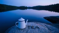 White enamel teapot on a granite rock by a calm lake at dusk, with the word TEA carved in stone and misty forest hills in the background.