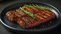 Juicy grilled beef steak with rosemary sprig and peppercorns on black plate. Medium rare meat with golden crust and grill marks shows professional cooking.