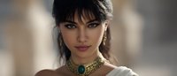 Young woman with dark hair wearing luxurious gold and emerald choker necklace and earrings against soft blurred background, elegant beauty portrait in warm tones.