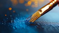 Artistic paintbrush with golden bristles touching glittery blue surface creating magical effect with bokeh lights in dark background for creative design projects.