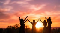 Four friends holding hands with raised arms celebrating together at sunset with dramatic orange and pink sky creating silhouettes against golden hour light.