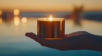 Glowing candle in glass holder on open palm against blurred sunset water background creates serene atmosphere for meditation and relaxation.