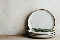 Minimalist ceramic plates stack with rosemary sprig on wooden shelf against neutral wall, modern kitchen tableware for dining and home decor styling.