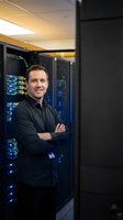 Confident IT professional standing with crossed arms in modern data center server room with glowing blue lights and network equipment for technology solutions.