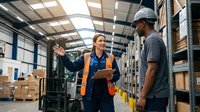 Female warehouse supervisor in safety vest discussing logistics with male worker in industrial storage facility with forklift and inventory shelves.