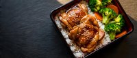Glazed chicken thighs with sesame seeds served over steamed rice alongside fresh broccoli and red bell peppers on a dark slate surface, suitable for food blogs and culinary publications.