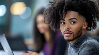Young African American professional with curly afro hairstyle wearing grey turtleneck sweater working at computer in modern office environment, looking away thoughtfully.