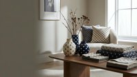 Neutral living room corner with wooden table displaying ceramic vases, dried flowers, patterned pillows, and stacked magazines near bright window for home decor and interior design projects.